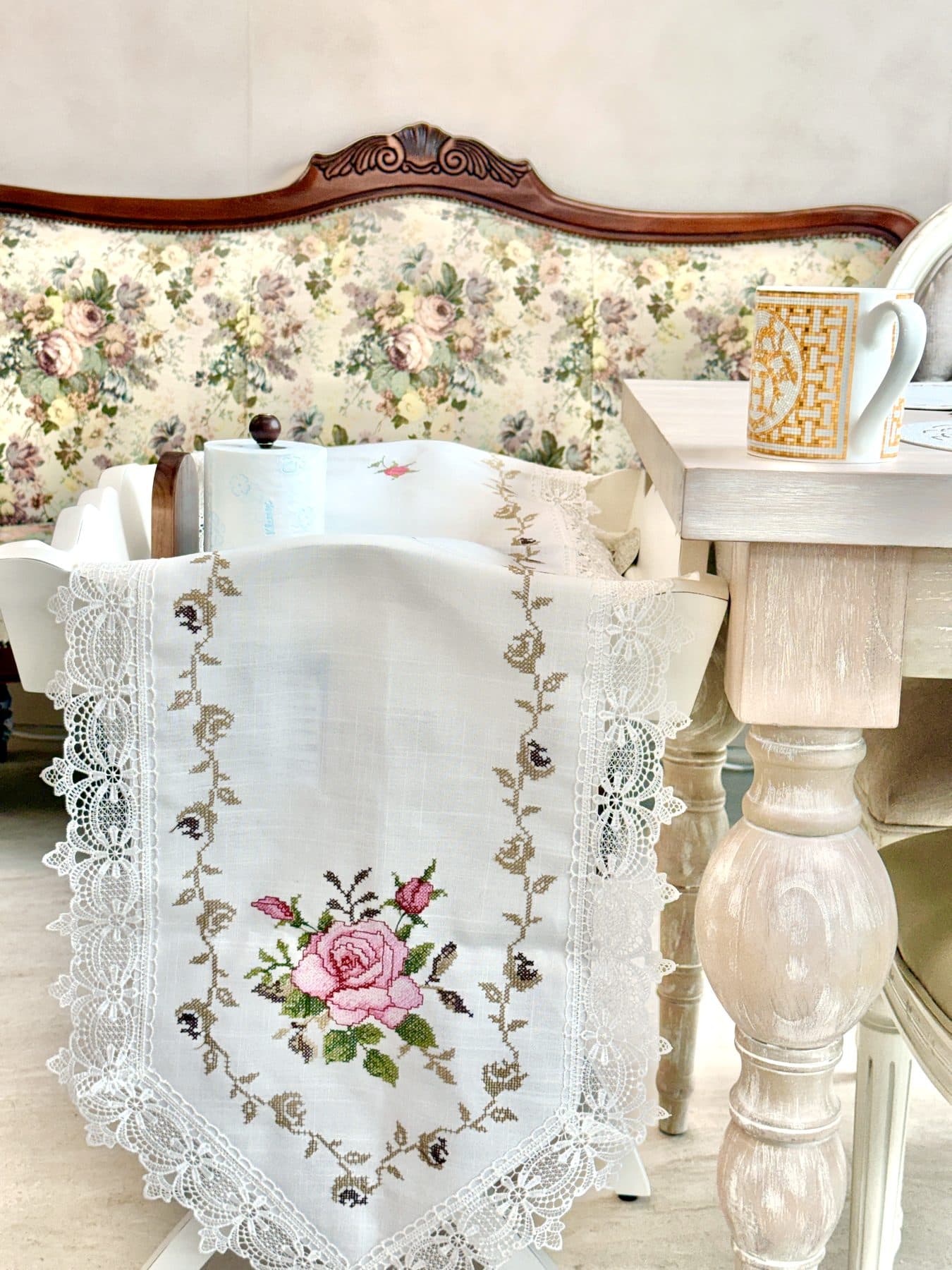 Un chemin de table en dentelle brodé d'une rose au point de croix, posé sur une chaise devant un canapé ancien à fleurs